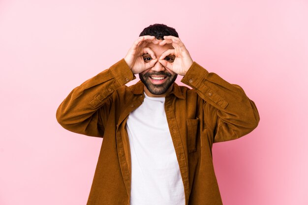 Young latin man against a pink wall isolated showing okay sign over eyes