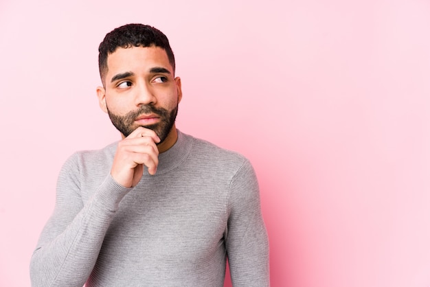 Young latin man against a pink wall looking sideways with doubtful and skeptical expression.