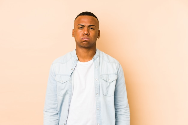 Young latin man on beige wall blows cheeks, has tired expression.