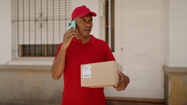 Young latin man delivery worker holding package talking on smartphone at street