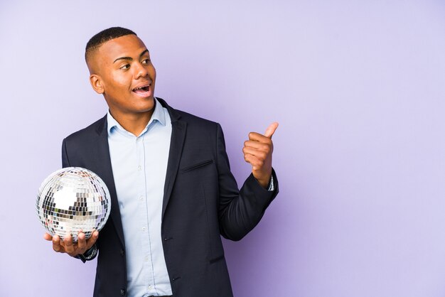 Young latin man holding a ball party points with thumb finger away, laughing and carefree.