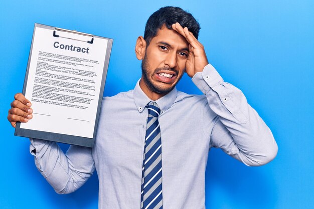 Young latin man holding clipboard with contract document stressed and frustrated with hand on head, surprised and angry face