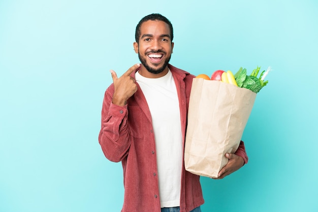 親指を立てるジェスチャーを与える青い背景で隔離の食料品の買い物袋を保持している若いラテン男性