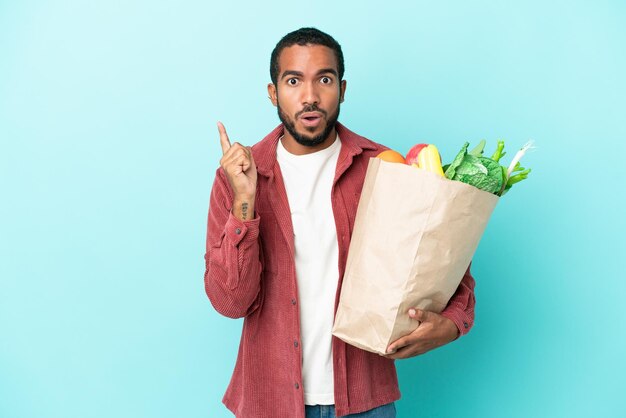 Молодой латиноамериканец, держащий продуктовую сумку на синем фоне, намереваясь реализовать решение, подняв палец вверх