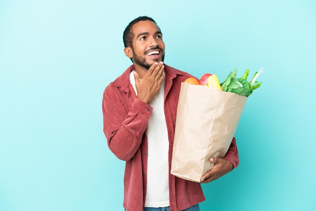 Young latin man holding a grocery shopping bag isolated on blue background looking up while smiling