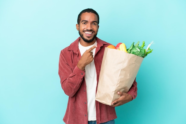 Young latin man holding a grocery shopping bag isolated on blue
background pointing to the side to present a product