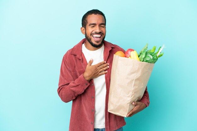 Young latin man holding a grocery shopping bag isolated on blue background smiling a lot