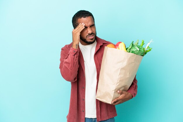 Young latin man holding a grocery shopping bag isolated on blue background with headache