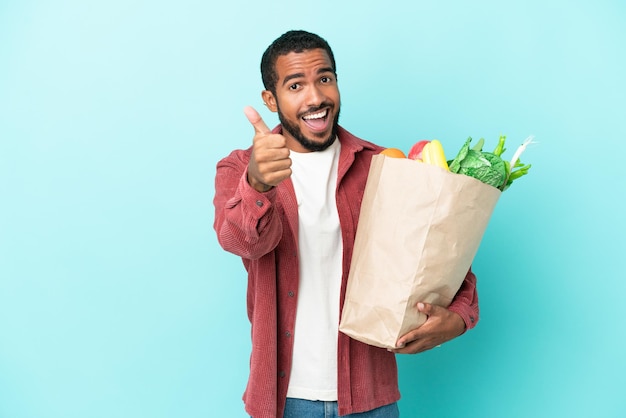 Young latin man holding a grocery shopping bag isolated on blue background with thumbs up because something good has happened