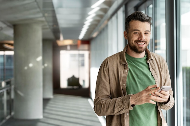 Young Latin man holding mobile phone standing in the office smiling Stylish guy using smartphone