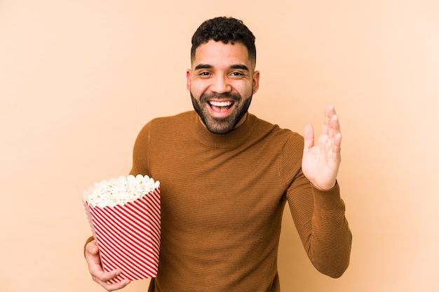 Young latin man holding a pop corn isolated receiving a pleasant surprise, excited and raising hands.