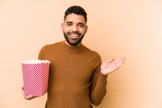 ポップコーンを持って孤立した若いラテン系男性は、手のひらにコピースペースを示し、腰に別の手を持っています。