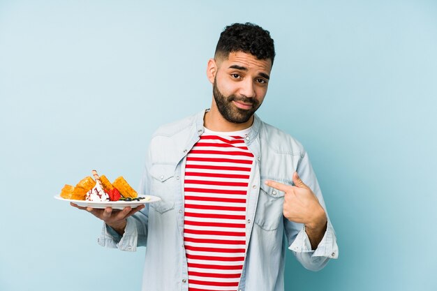 Young latin man holding a waffle isolated pointing with finger at you as if inviting come closer.