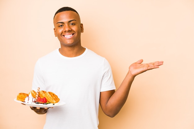 Young latin man holding a waffle isolated showing a copy space on a palm and holding another hand on waist.