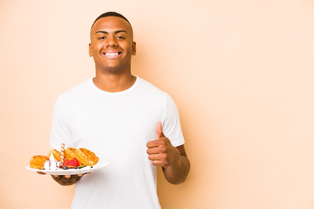 Young latin man holding a waffle isolated smiling and raising thumb up