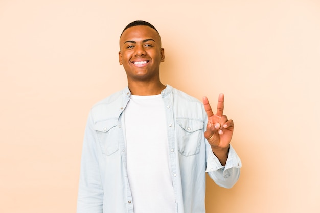 Young latin man isolated on beige wall showing victory sign and smiling broadly.