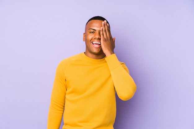 Young latin man isolated on purple wall having fun covering half of face with palm