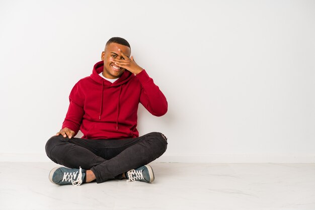 Young latin man sitting on the floor isolated blink at the camera through fingers, embarrassed covering face.