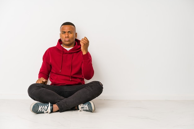 Young latin man sitting on the floor isolated showing fist, aggressive facial expression.