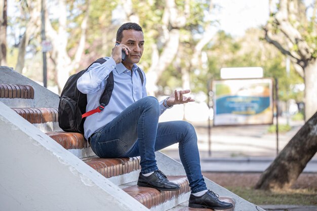 Young latin man sitting on a square bench using his mobile phone