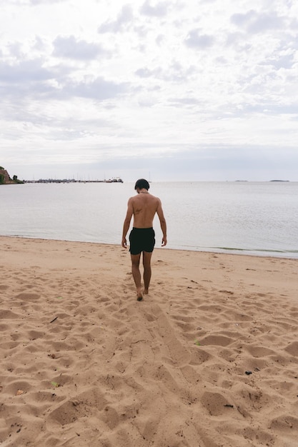 Young latin man in swimwear walking on the sand while watching his footsteps Vertical photo