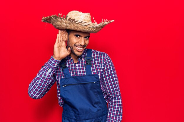 Молодой латиноамериканец в фермерской шляпе и фартуке улыбается, прикрывая ухо рукой, прислушиваясь к слухам или сплетням о концепции глухоты