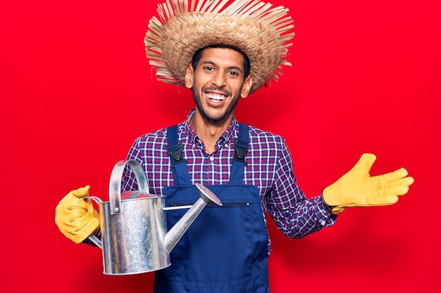 Young latin man wearing farmer hat and gloves holding watering can celebrating achievement with happy smile and winner expression with raised hand