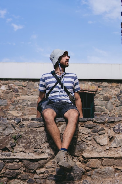 Young latin man with a bucket hat sitting on a stone wall looking to the side Vertical photo