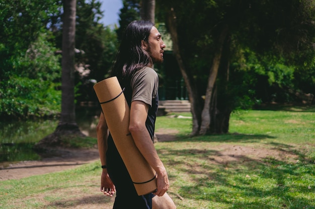 Photo young latin man with long hair walking in a green area with a yoga mat on his back