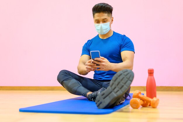 Young Latin man with protective face mask, relaxed, using mobile phone after workout in gym during pandemic new normal.