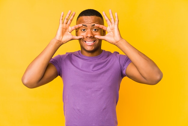 Young latin man on yellow wall keeping eyes opened to find a success opportunity.