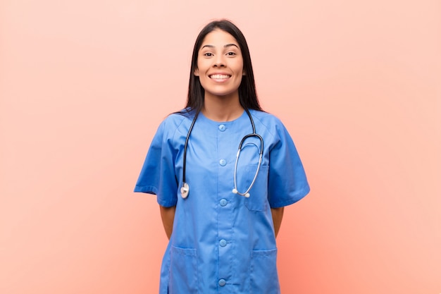 Фото Молодая латинская медсестра выглядит счастливой и тупой с широкой, веселой, чокнутой улыбкой и широко открытыми глазами на розовой стене