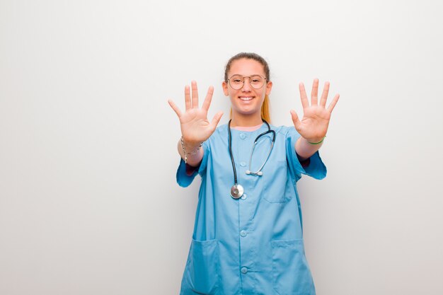 Young latin nurse smiling and looking friendly, showing number ten or tenth with hand forward, counting down against white wall