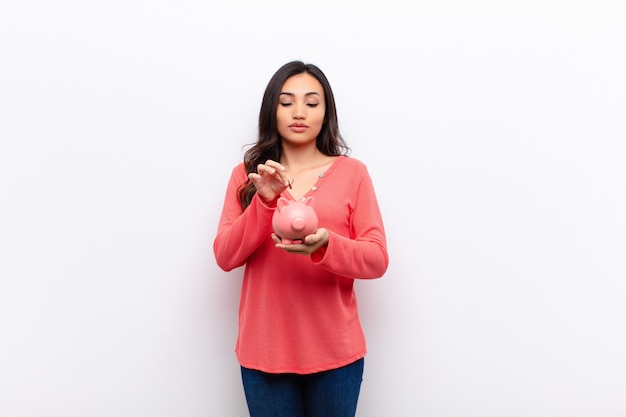 Young latin pretty woman  against flat wall with a piggy bank