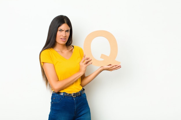 Young latin pretty woman confused, doubtful, thinking, holding the letter Q of the alphabet to form a word or a sentence