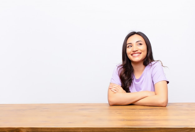 Young latin pretty woman feeling happy, proud and hopeful, wondering or thinking, looking up to copy space with crossed arms against flat wall