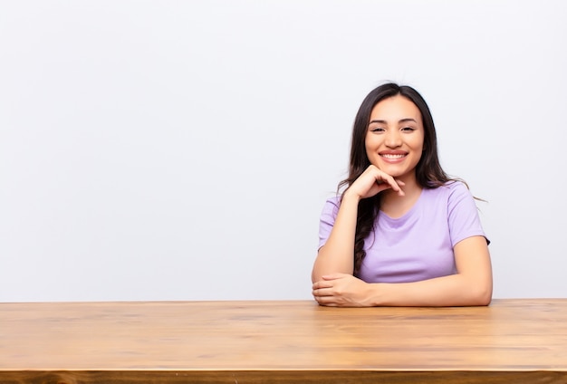 Молодая латинская симпатичная женщина, выглядящая счастливой и улыбающаяся с рукой на подбородке, задающаяся вопросом или задающая вопрос, сравнивая варианты с плоской стеной