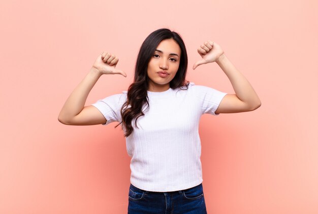 Young latin pretty woman looking sad, disappointed or angry, showing thumbs down in disagreement, feeling frustrated against flat wall