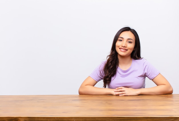 Young latin pretty woman smiling happily with a hand on hip and confident, positive, proud and friendly attitude against flat wall