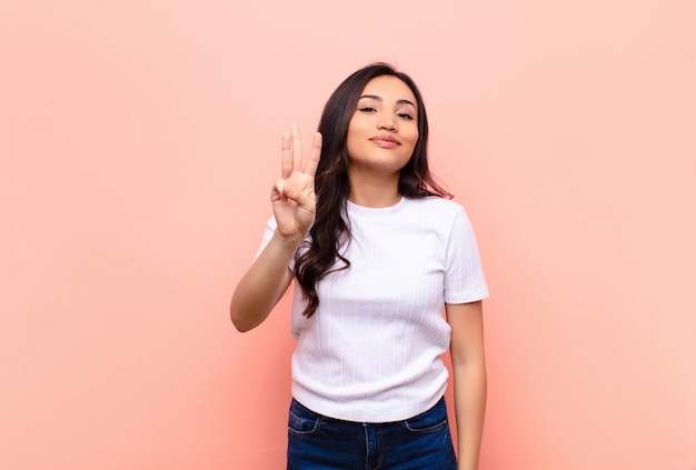 Young latin pretty woman smiling and looking friendly, showing number three or third with hand forward, counting down against flat wall