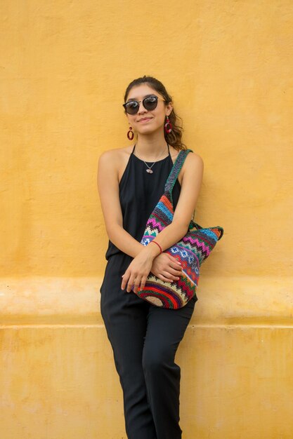 Young latin tourist in black jumpsuit using a mexican handmade\
bag. yellow wall. vertical photo