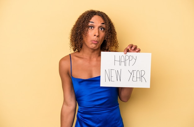 黄色の背景で隔離された新年を祝う若いラテン系の性転換者の女性は肩をすくめると混乱した目を開きます。