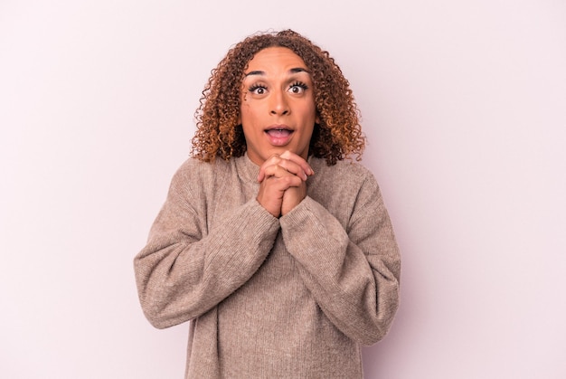 Young latin transsexual woman isolated on pink background praying for luck, amazed and opening mouth looking to front.