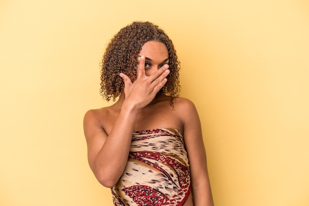 黄色の背景で隔離された若いラテン系の性転換者の女性は、恥ずかしい顔を覆って、指を通してカメラで点滅します。