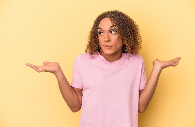 Giovane donna transessuale latina isolata su sfondo giallo confusa e dubbiosa che scrolla le spalle per tenere uno spazio di copia.