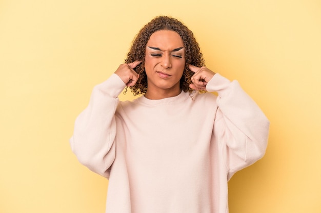 노란색 배경에 고립된 젊은 라틴 성전환 여성은 큰 소리로 스트레스를 받고 절망적으로 귀를 덮고 있습니다.