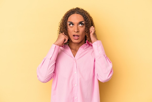 指で耳を覆っている黄色の背景に隔離された若いラテン系トランスセクシュアル女性は、大声で周囲にストレスと絶望的です。