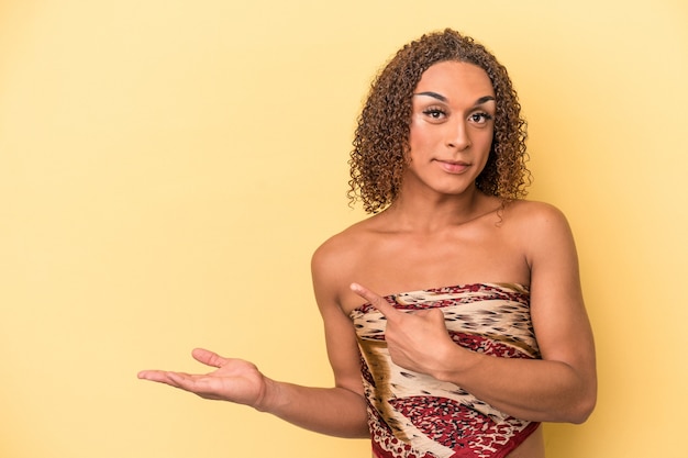 Young latin transsexual woman isolated on yellow background excited holding a copy space on palm.