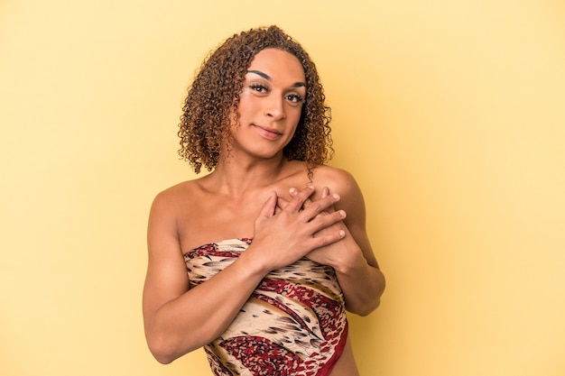 Young latin transsexual woman isolated on yellow background has friendly expression, pressing palm to chest. Love concept.