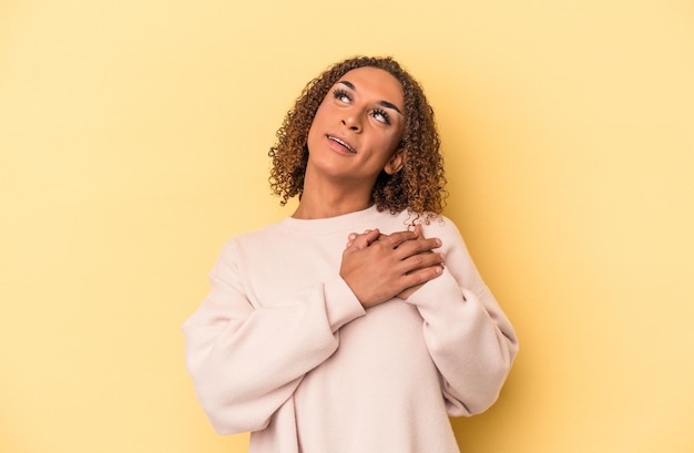 Young latin transsexual woman isolated on yellow background has friendly expression, pressing palm to chest. Love concept.